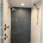 Dark grey herringbone patterned tiles forming an accent wall inside a marble shower stall.