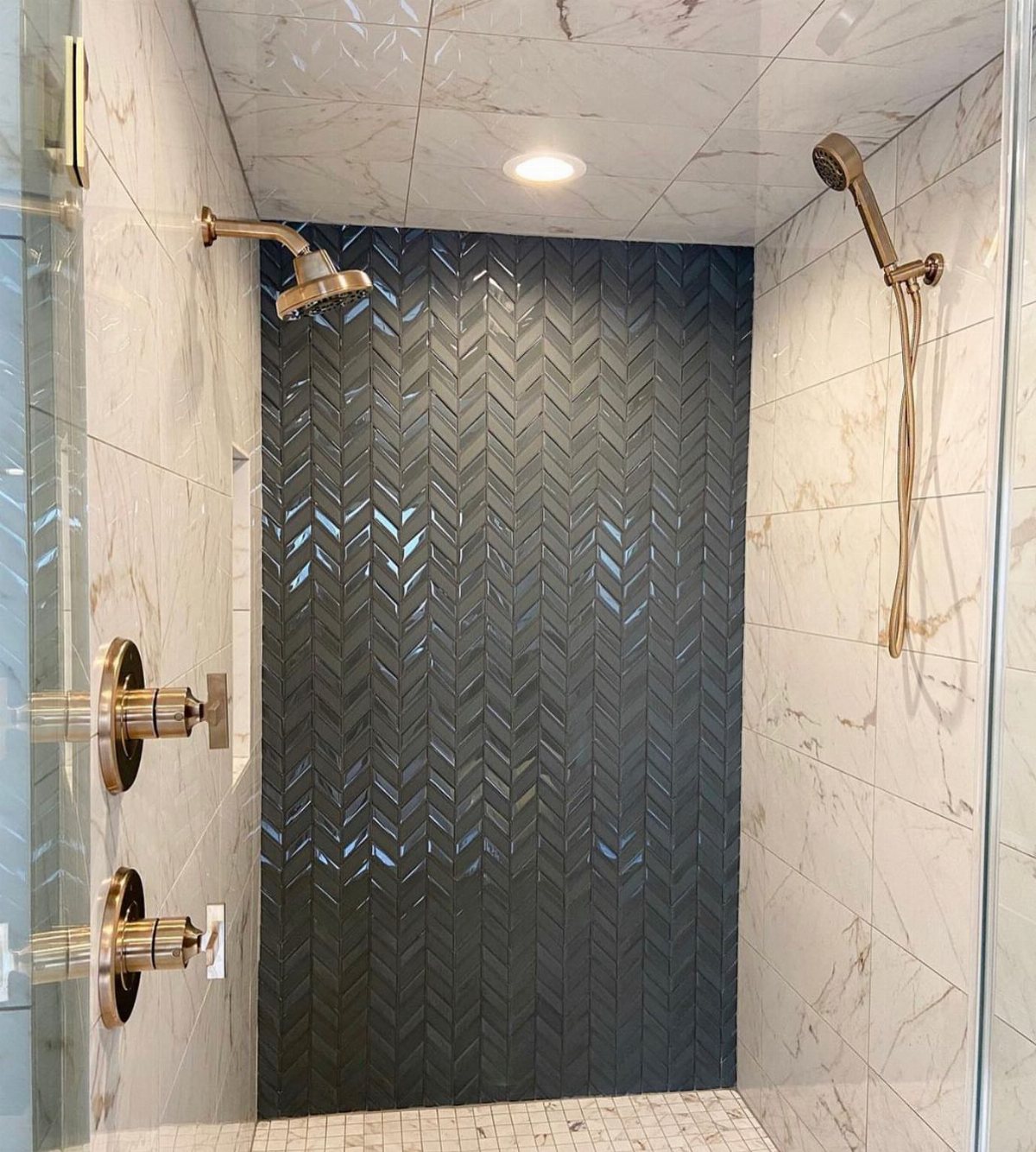 Dark grey herringbone patterned tiles forming an accent wall inside a marble shower stall.